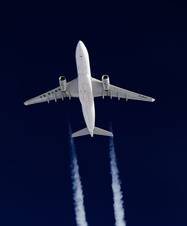 A white airplane is flying overhead against a deep blue sky, leaving two white contrails behind it. The aircraft is viewed from below, clearly showing its wings and engines.
