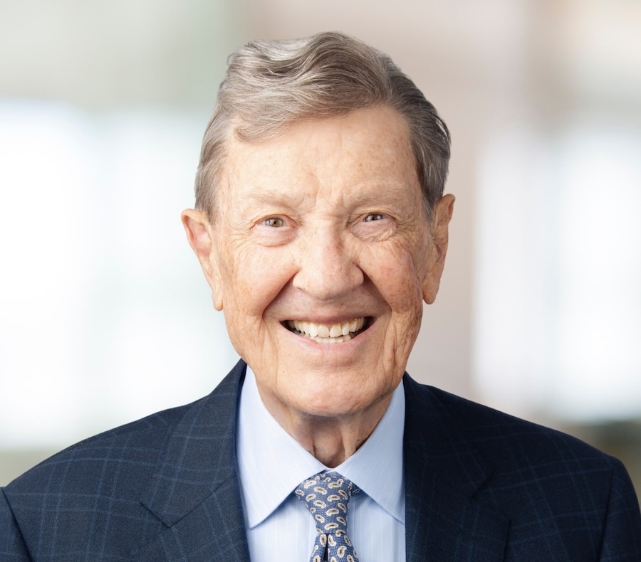 An elderly man with gray hair smiles warmly, wearing a dark checkered suit, a light blue shirt, and a patterned tie. The background is softly blurred with light tones.