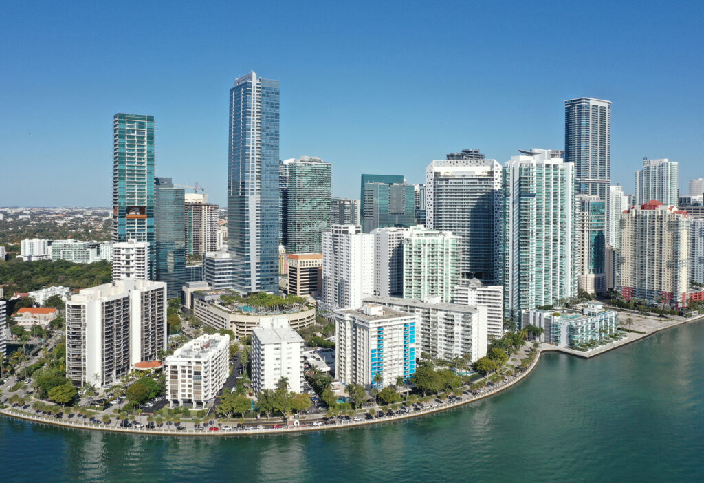Aerial view of a modern city skyline with high-rise buildings along a waterfront, clear blue sky, and green trees lining the shore.
