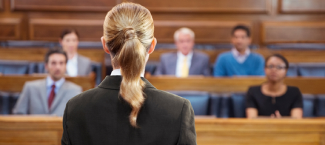 A woman with blonde hair in a ponytail stands in a courtroom, facing a seated jury of four men and one woman, with wooden benches and a judge’s bench visible in the background.