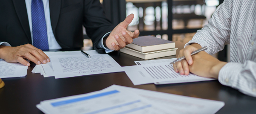 Two people in business attire sit at a table discussing documents, with one gesturing and the other taking notes. Papers, a pen, and closed books are spread out on the table.