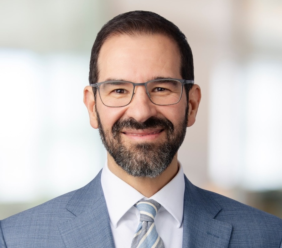 A man with short dark hair, glasses, and a beard is smiling. He is wearing a light blue suit jacket, a white dress shirt, and a light striped tie, with a blurred background behind him.