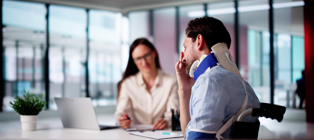 A man wearing a neck brace and arm sling sits across from a woman at a desk in an office, looking distressed. The woman appears to be taking notes or discussing something with him.