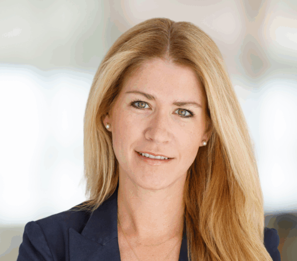 A woman with long straight blonde hair, wearing a navy blue blazer smiles at the camera against a softly blurred indoor background.