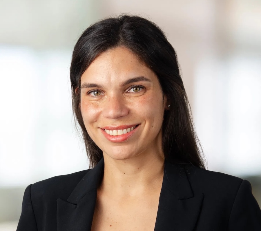 A woman with long dark hair, wearing a black blazer over a red top, smiles at the camera against a softly blurred indoor background.