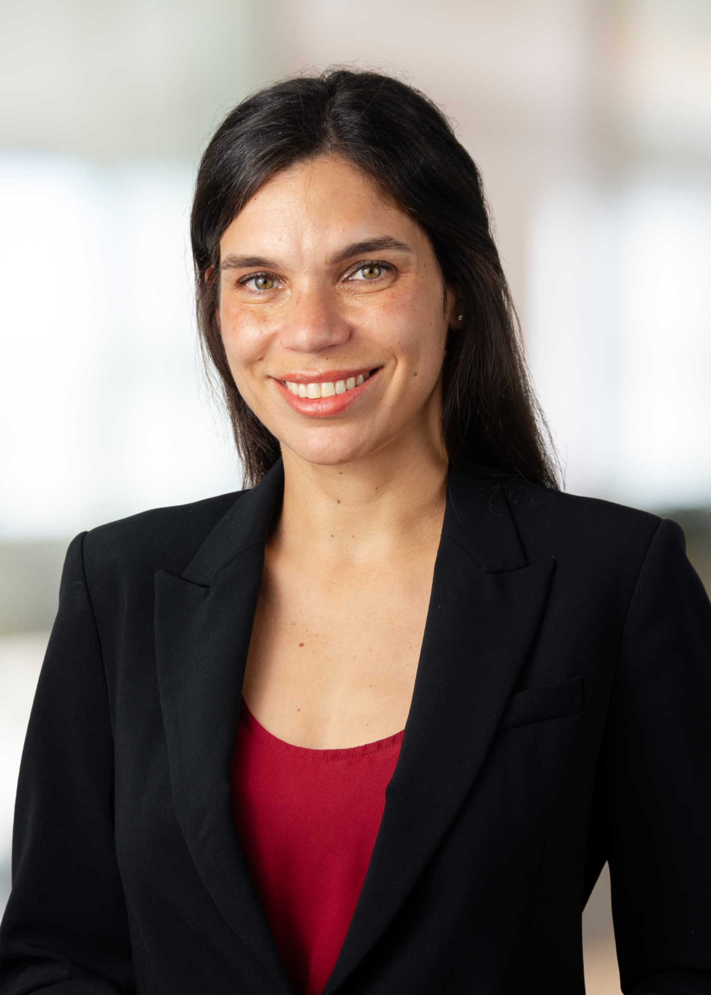 A woman with long dark hair, wearing a black blazer over a red top, smiles at the camera against a softly blurred indoor background.