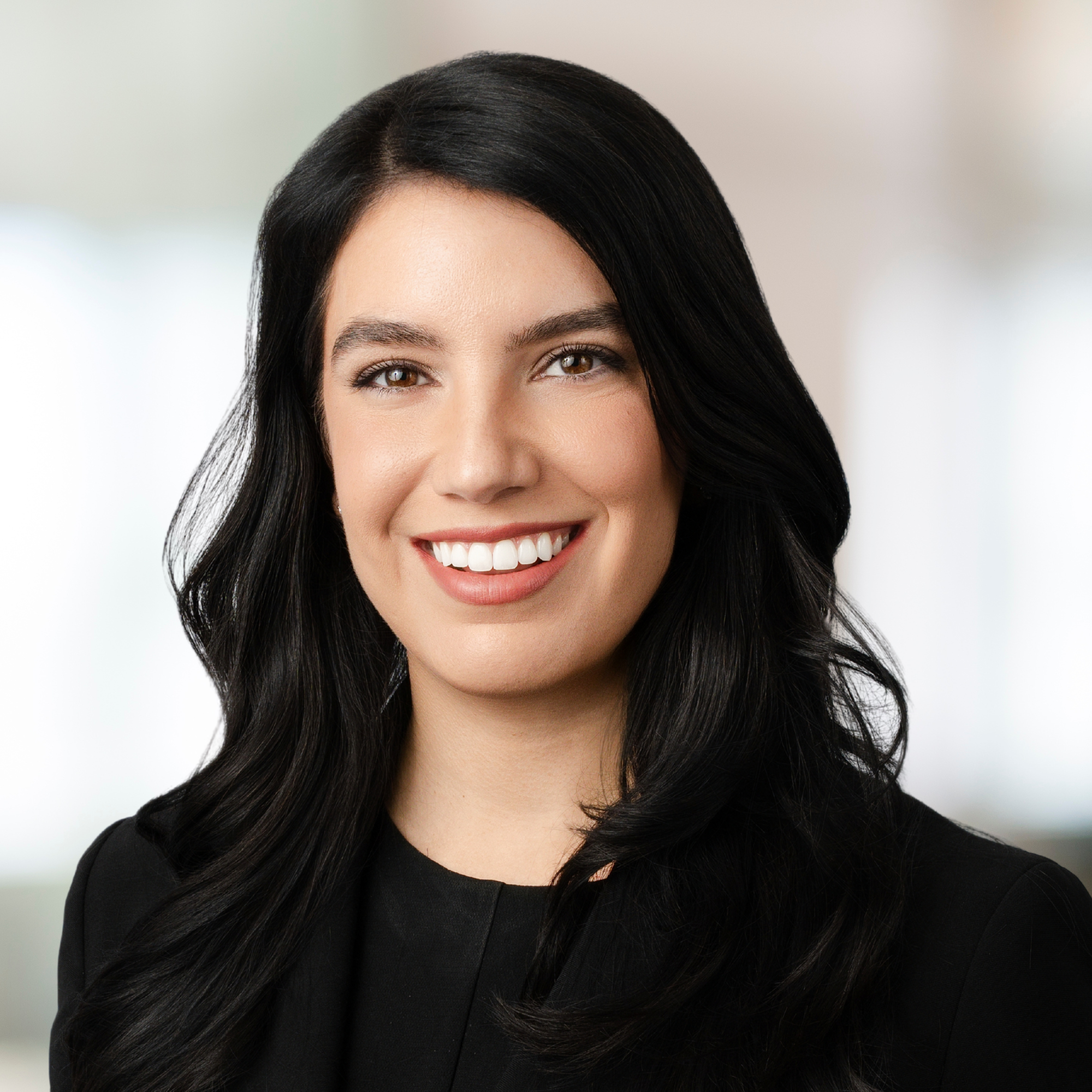 A professional headshot of a woman with long, dark, wavy hair, smiling at the camera, wearing a black blazer.