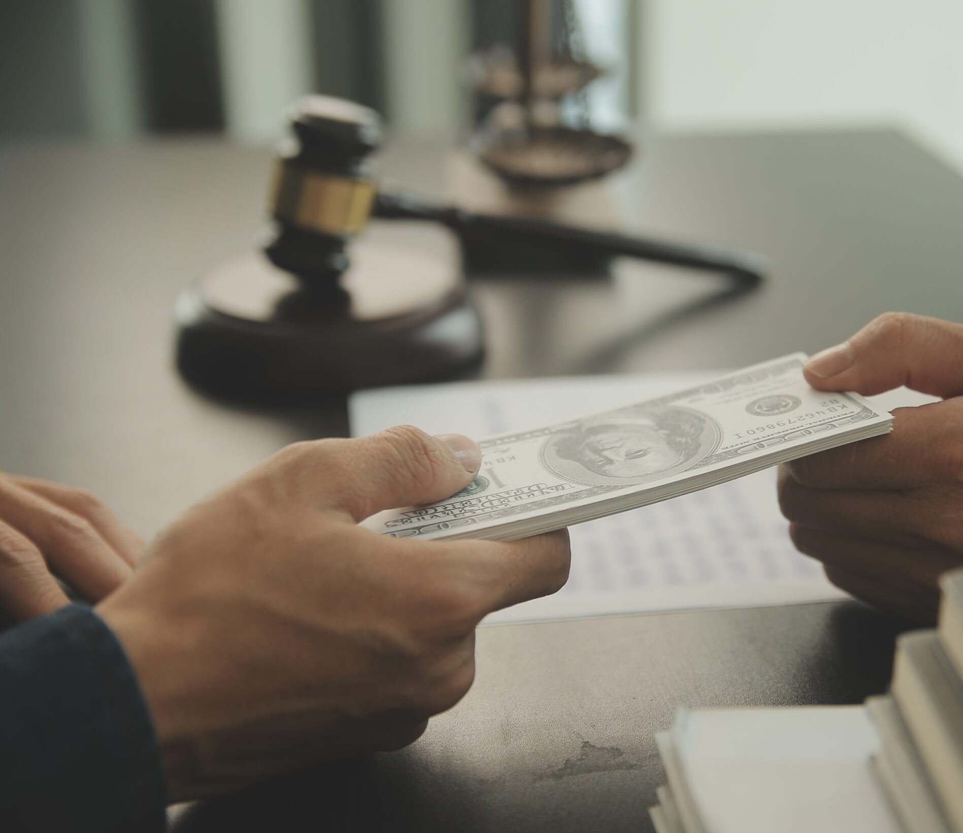 A person hands a stack of U.S. hundred-dollar bills to another person at a desk, with a gavel, legal documents, and a pen visible, suggesting a legal or financial transaction.