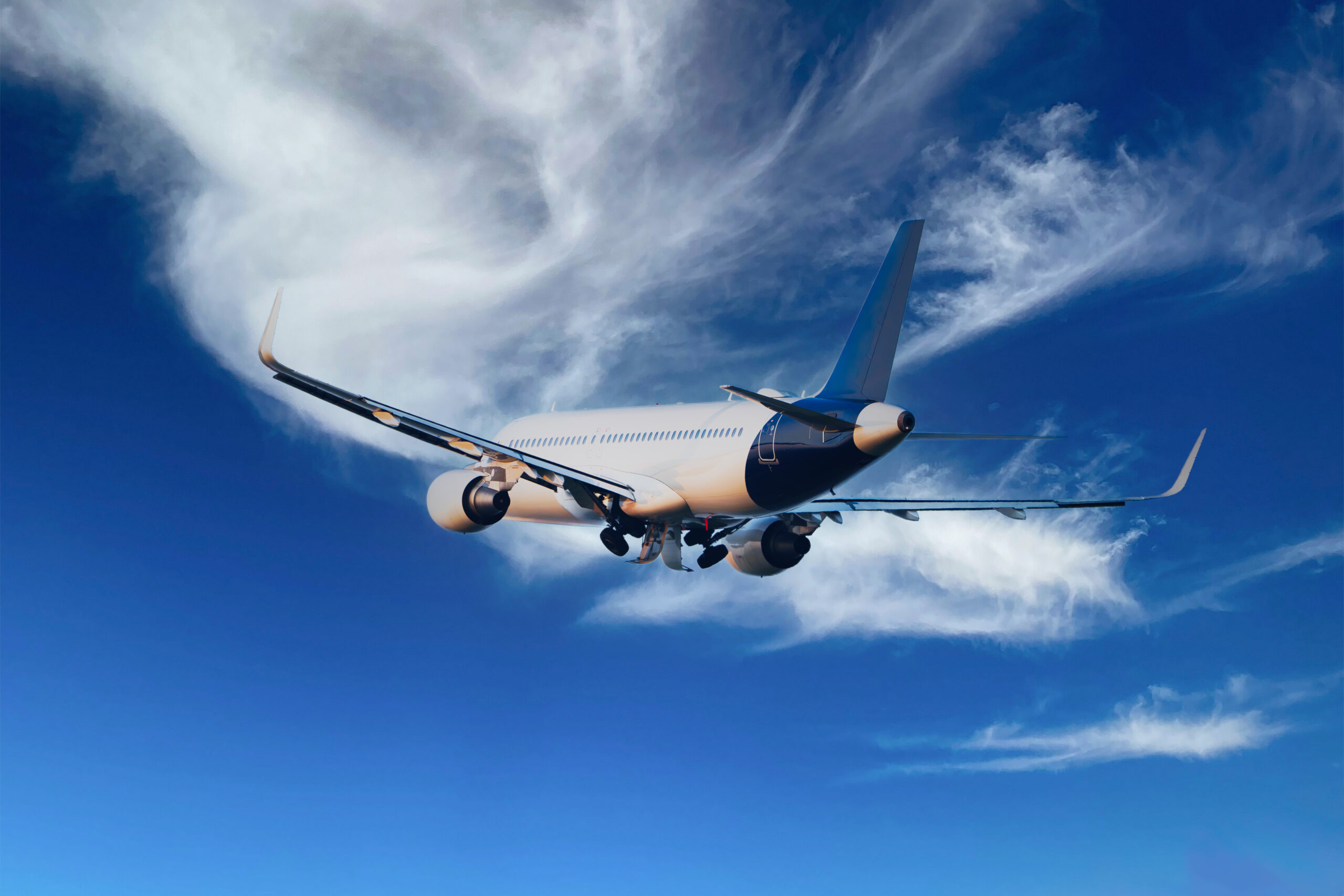 A commercial airplane flies through a clear blue sky with wispy white clouds, captured from behind and slightly below the aircraft.