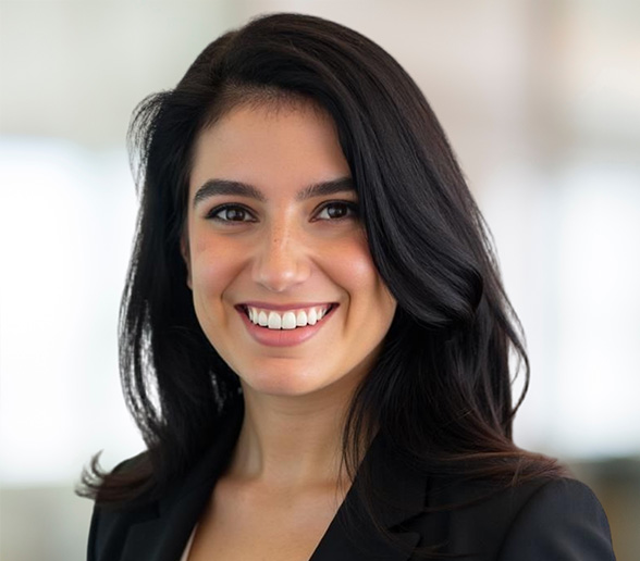 A woman with long, dark hair and a bright smile is wearing a black blazer. She is looking at the camera with a blurred, light-colored background behind her.