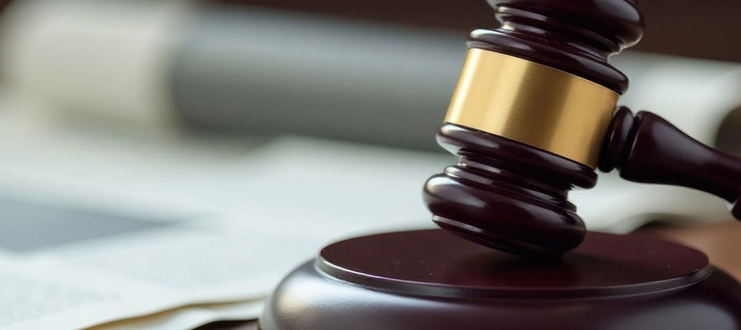 A close-up of a wooden judge’s gavel with a gold band resting on a sound block, with blurred documents in the background.