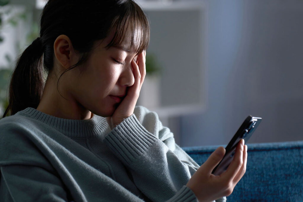 A woman sits indoors, holding a smartphone in one hand while covering her face with the other, appearing distressed or upset. She is wearing a light sweater and has her hair tied back.