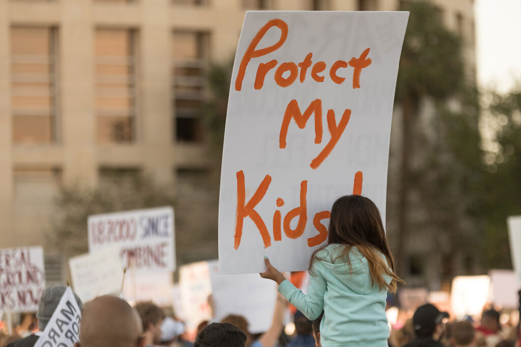 A young girl at a protest holds a large sign that reads Protect My Kids! in bold orange letters. Other protestors and signs are visible in the background.