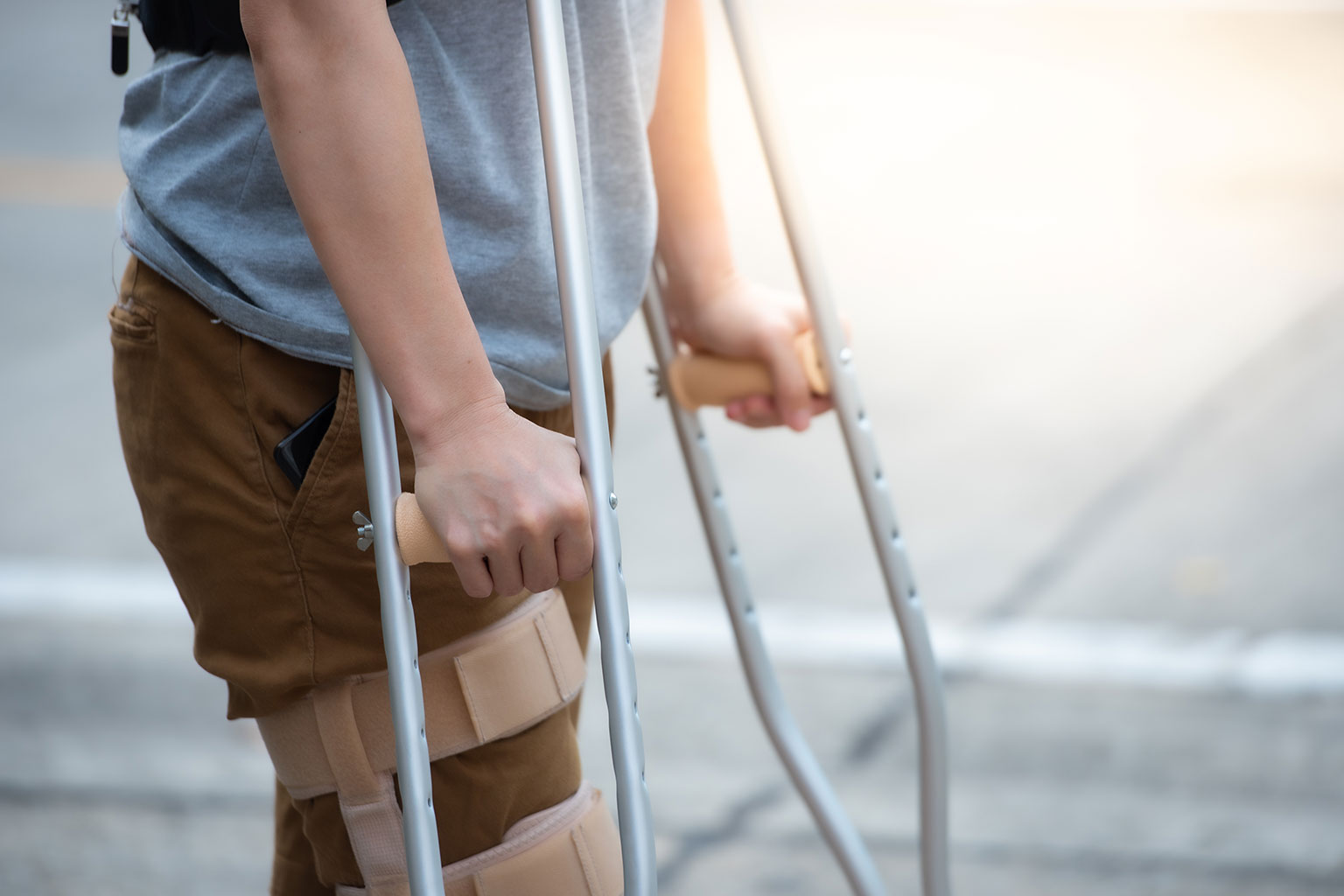 A person wearing brown pants and a gray shirt uses crutches, with both knees wrapped in supportive braces, standing on a paved surface outdoors.