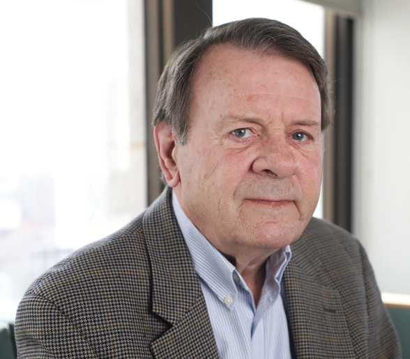 An older man with short brown hair is wearing a checked blazer and light blue shirt, sitting indoors in front of large windows with a neutral expression.