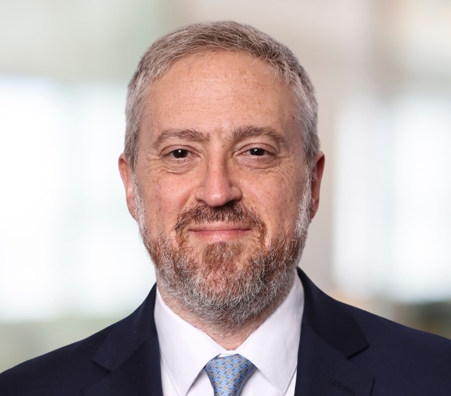 A middle-aged man with short gray hair and a beard, wearing a dark suit, white shirt, and light blue patterned tie, is smiling softly in a brightly lit, blurred indoor setting.