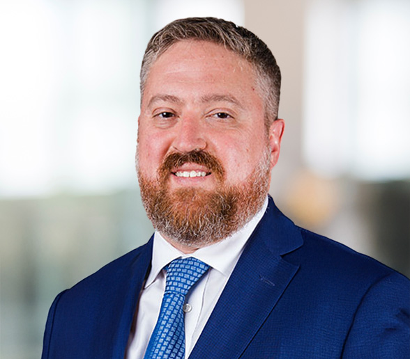 A man with short brown hair and a beard, wearing a blue suit, white shirt, and blue patterned tie, smiles at the camera against a blurred indoor background.