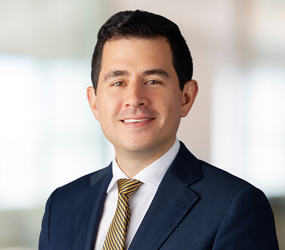 A man in a dark suit, white shirt, and patterned yellow tie smiles at the camera. He has short dark hair and is standing in front of a softly blurred background.