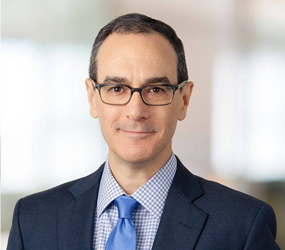 A man wearing glasses, a dark suit, a blue tie, and a checkered shirt poses for a professional headshot with a blurred office background.