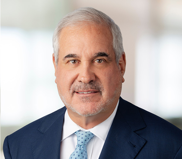 Attorney Steven C. Marks, a partner at Podhurst Orseck, wearing a blue suit, white shirt, and light blue polka dot tie, poses against a blurred indoor background.