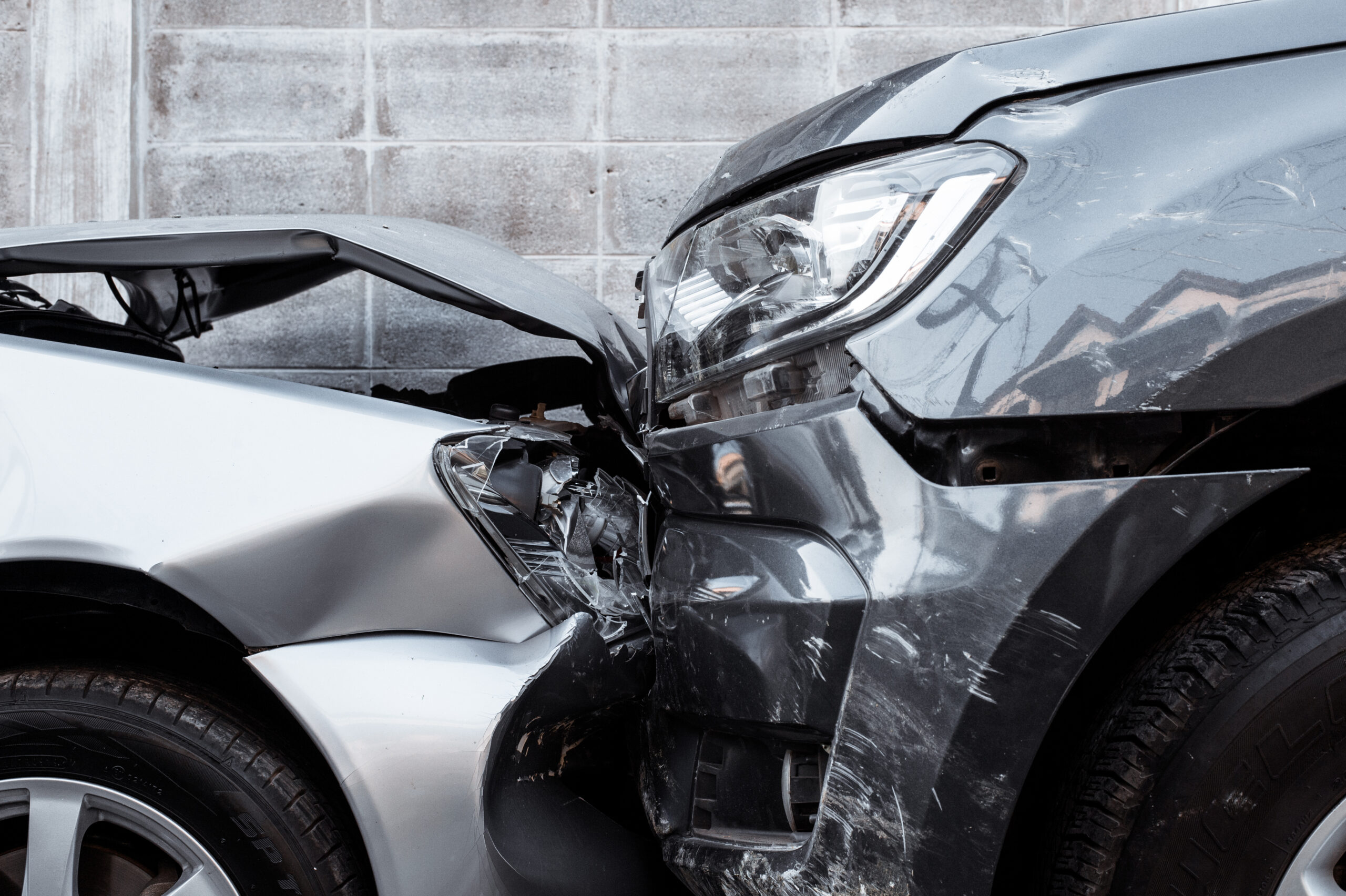 Two cars involved in a head-on collision, with visible damage to the front ends of both vehicles, including crumpled hoods and broken headlights, against a gray concrete wall.