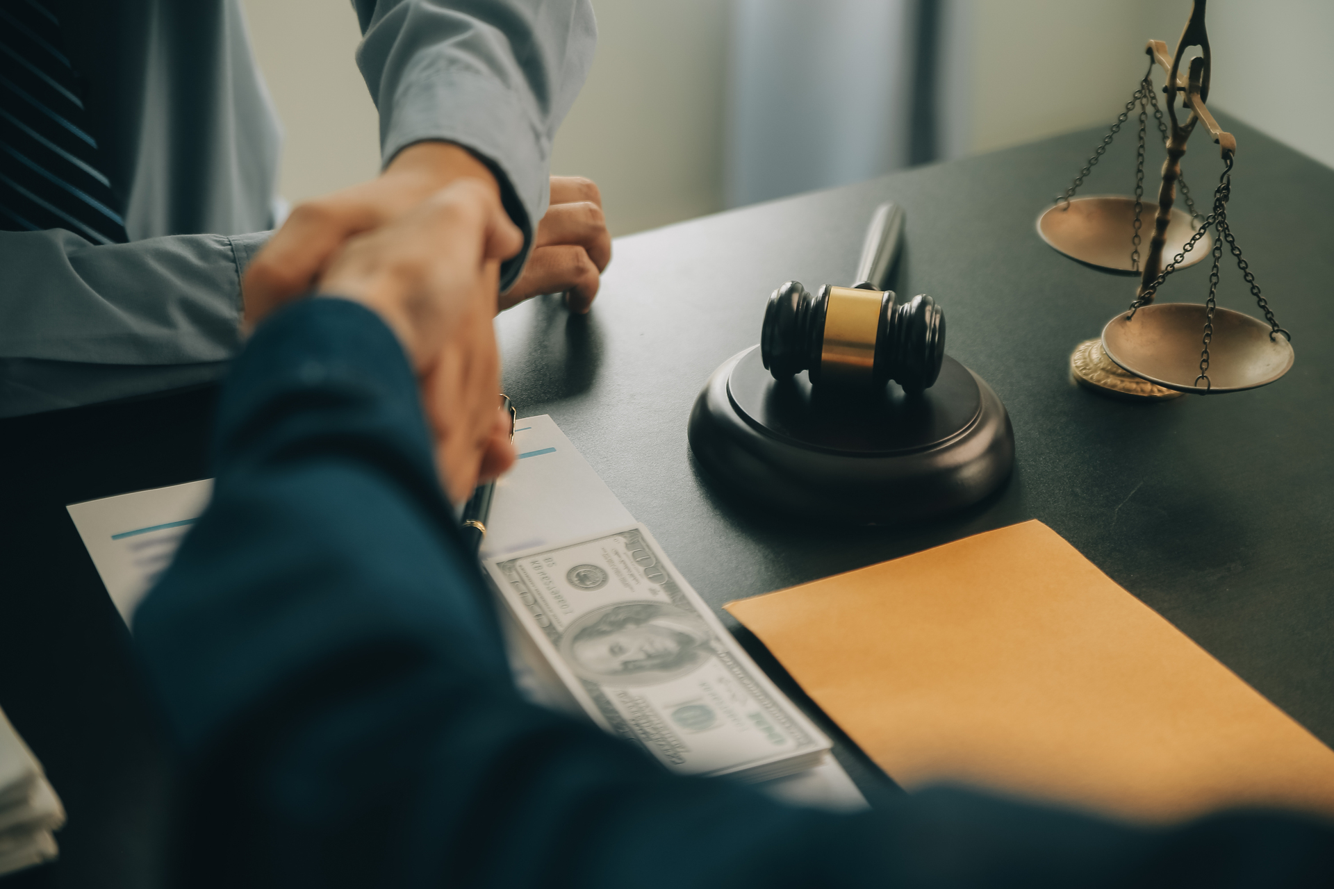 Two people shake hands over a desk with a gavel, scales of justice, an envelope, and a contract with hundred-dollar bills, suggesting a legal or financial agreement being made.