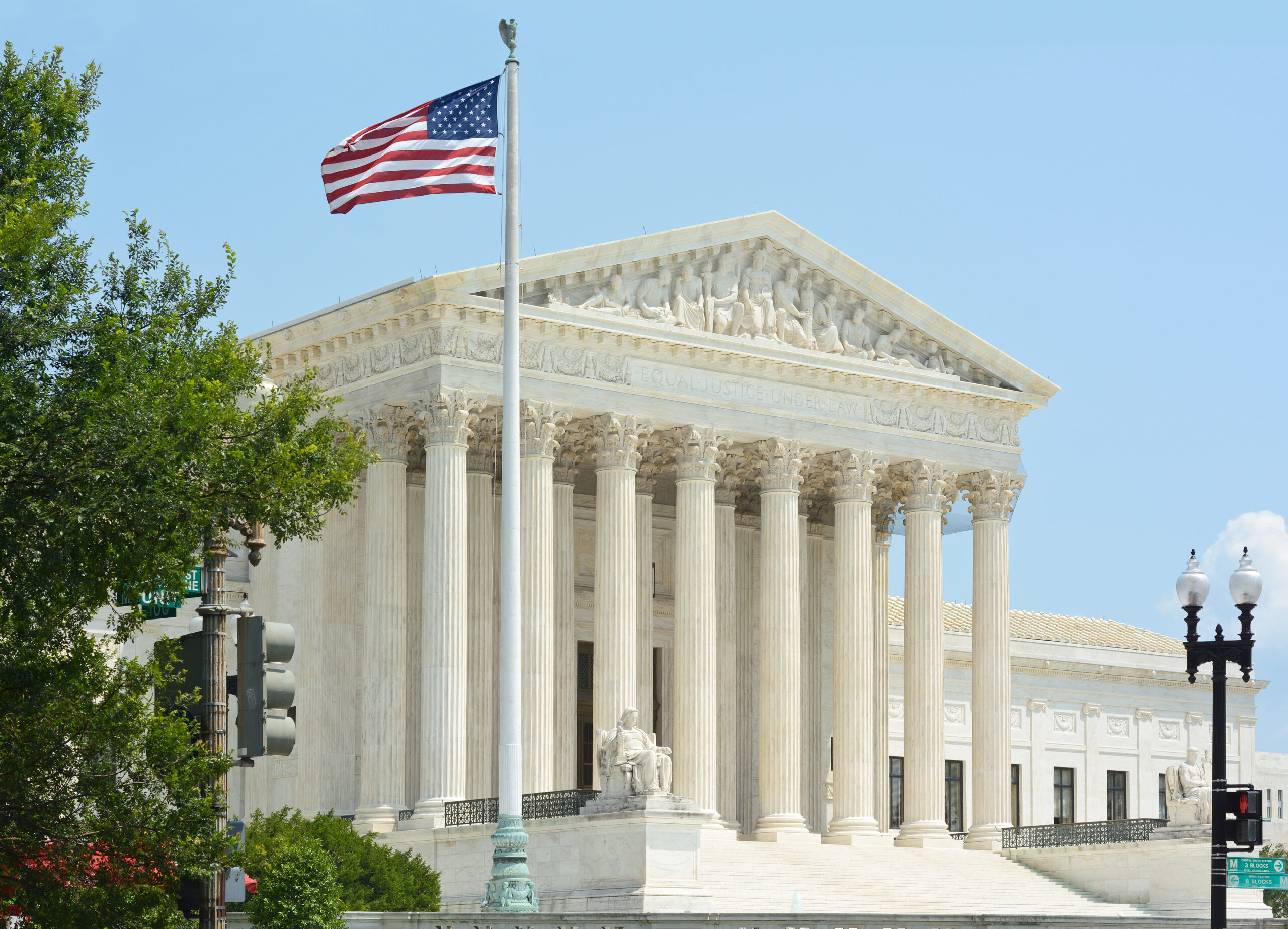 The United States Supreme Court building in Washington, D.C., with large white columns, intricate sculptures, and an American flag waving on a flagpole in the foreground under a clear blue sky.