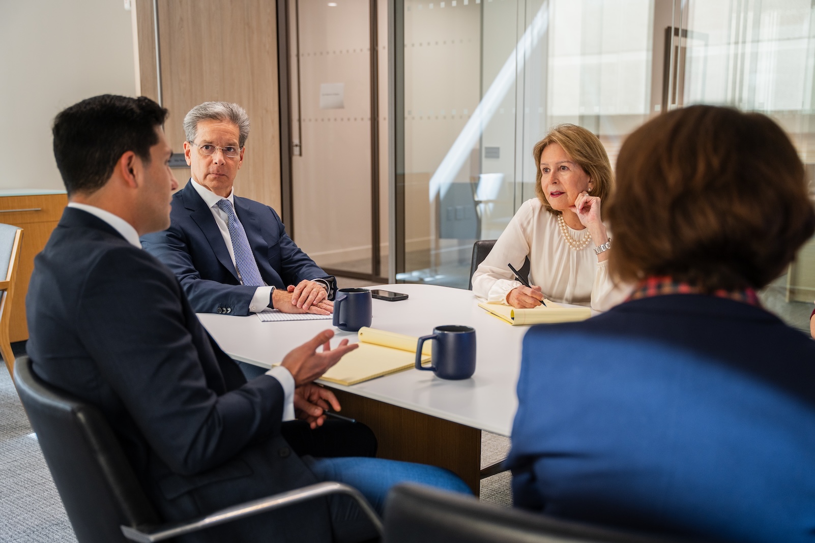 Four professionals in business attire sit around a table in a modern office, engaged in a serious discussion. Notebooks and mugs are on the table, and one person gestures while others listen attentively.