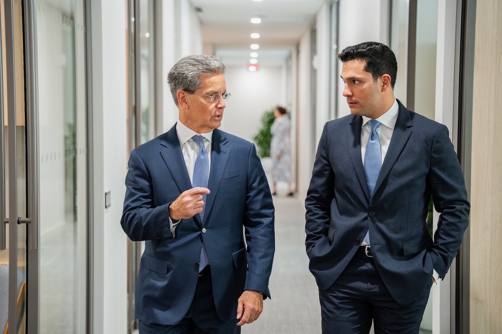 Two men in business suits walk down an office hallway, engaged in conversation. Both appear focused and professional. The background shows a bright corridor with glass walls and another person blurred in the distance.