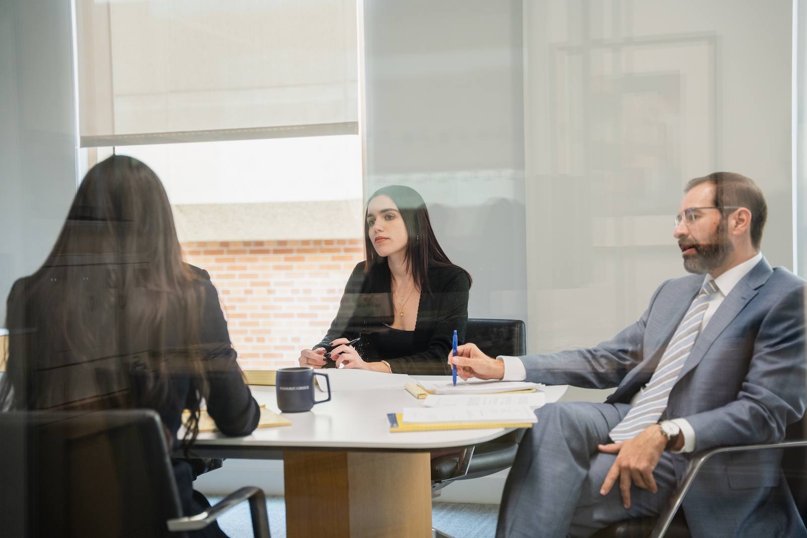 Three people in business attire sit around a meeting table in an office, two facing one. They appear to be in discussion, with notebooks, pens, and a coffee mug on the table. The image is taken through glass.