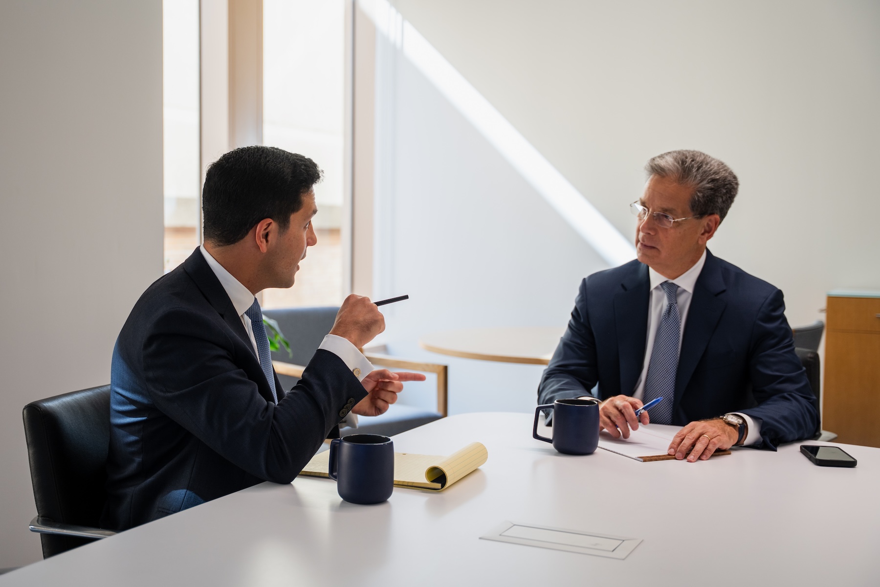 Two men in business suits sit at a table in a bright office, having a serious discussion. Both have notebooks and coffee mugs in front of them, and one gestures with a pen while speaking to the other.