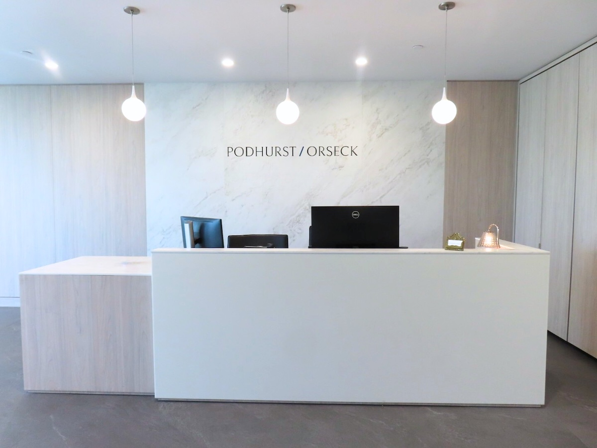 A modern, minimalist office reception area with a white desk, two computer monitors, three hanging pendant lights, and PODHURST / ORSECK on the marble-patterned back wall.