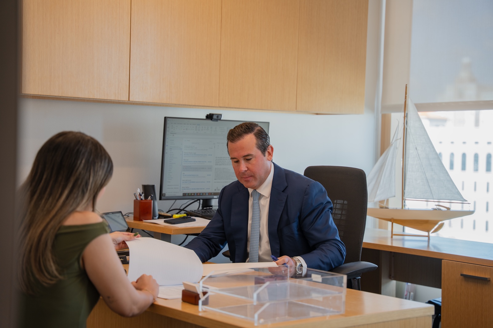 A man in a suit sits at a desk in a modern office, looking at documents with a woman across from him. A computer, office supplies, and a model sailboat are visible in the background.