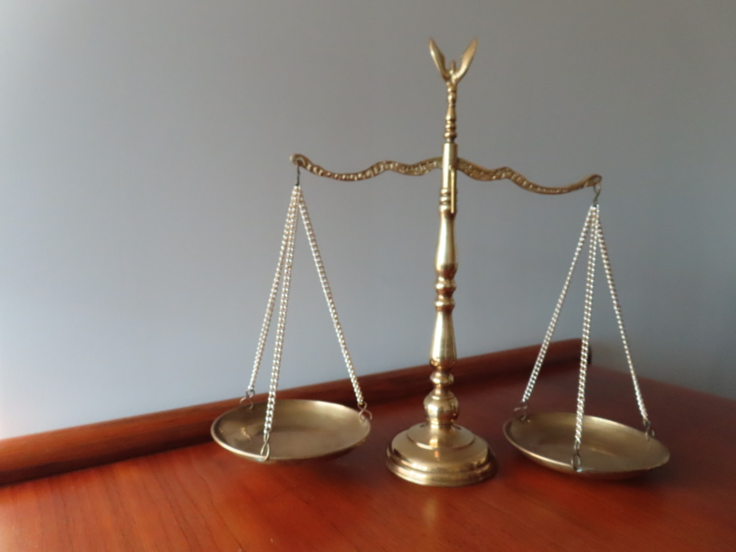 A brass balance scale with two unevenly balanced plates sits on a wooden surface against a plain light-colored wall.