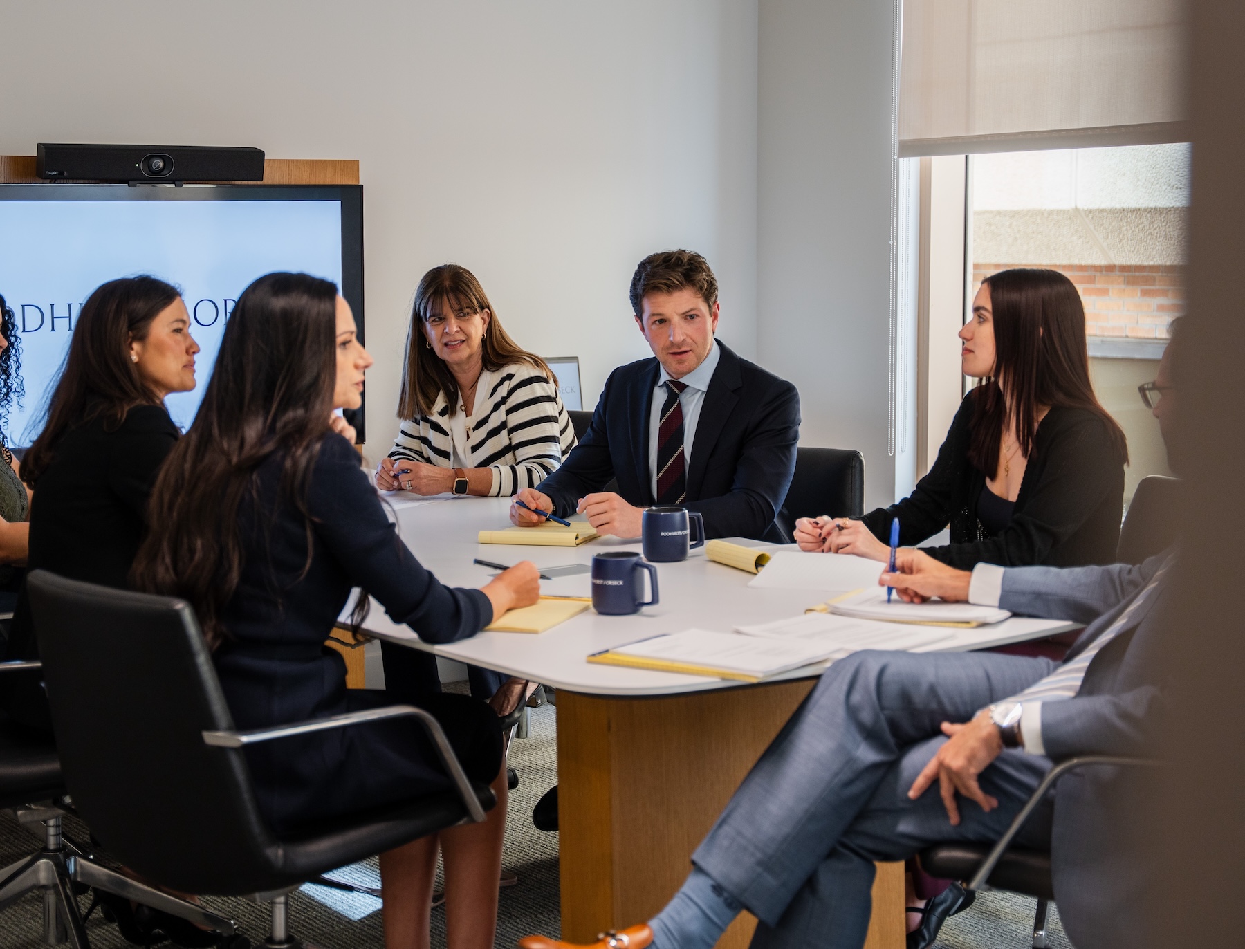 Modern office reception area with a receptionist sitting at a desk, minimalist decor, wooden chairs, a sofa, large windows providing natural light, and PODHURST/ORSECK written on the wall behind the desk.
