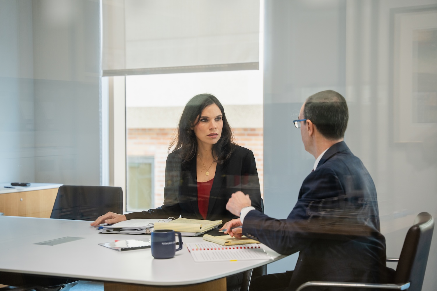 Two people in business attire sit at a table in a modern office, engaged in a serious conversation. Papers, a notebook, and a mug are on the table. The scene is viewed through a glass wall.