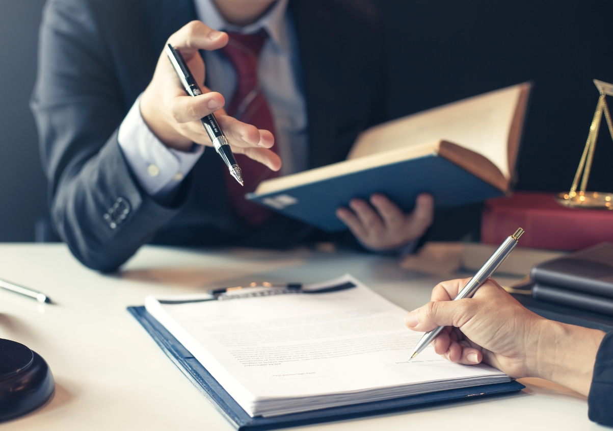 Two people in business attire sit at a desk discussing documents. One person gestures with a pen and holds an open book, while the other writes on a clipboard. A gavel and scales of justice are visible on the desk.