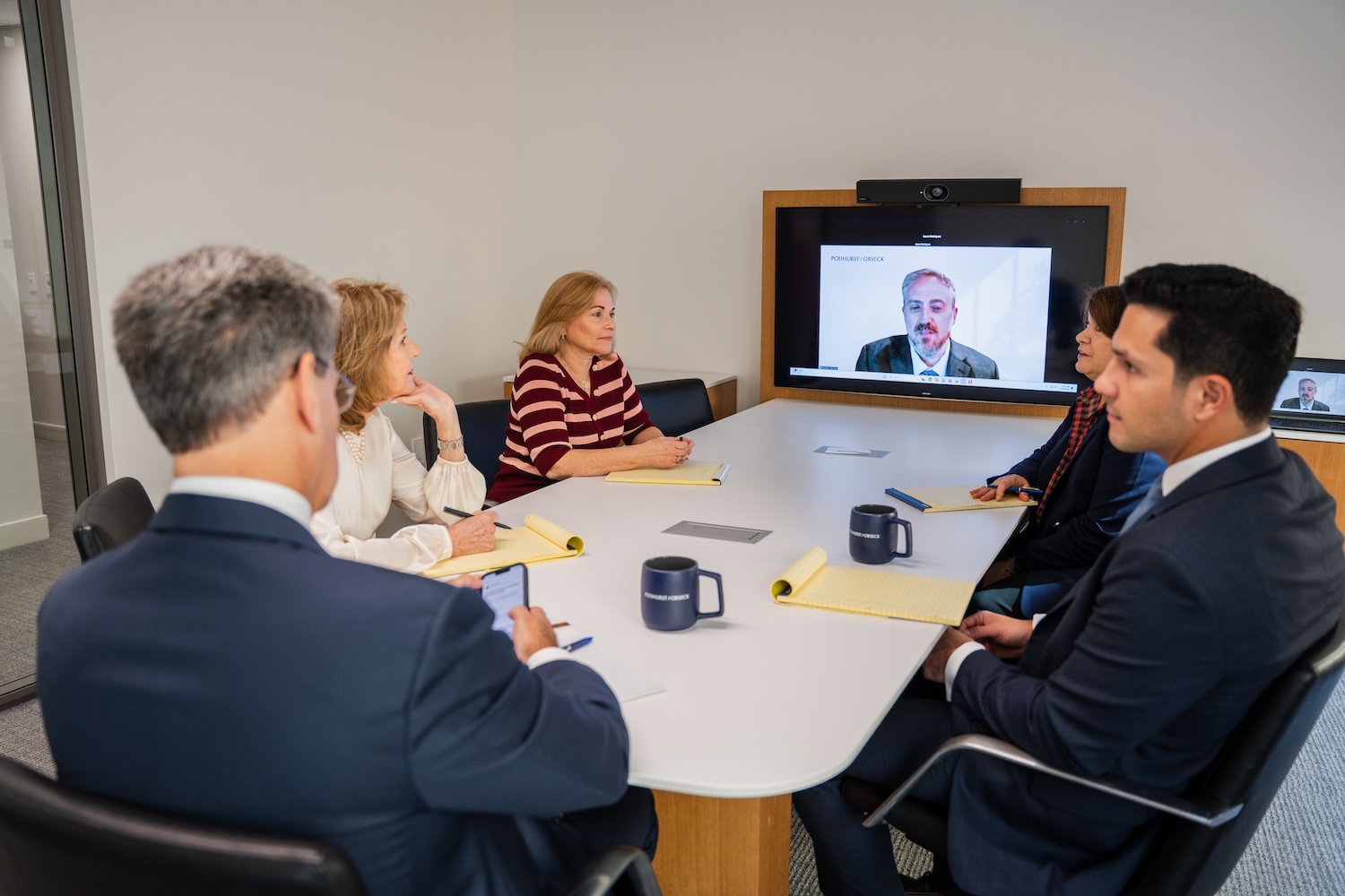 Five people sit around a conference table with notebooks and coffee mugs, while a sixth person joins the meeting via video call on a screen at the end of the table.