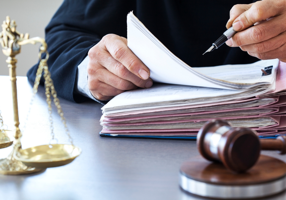 A person in formal attire reviews documents at a desk with a gavel, a stack of folders, and a brass balance scale, symbolizing legal work or a courtroom setting.