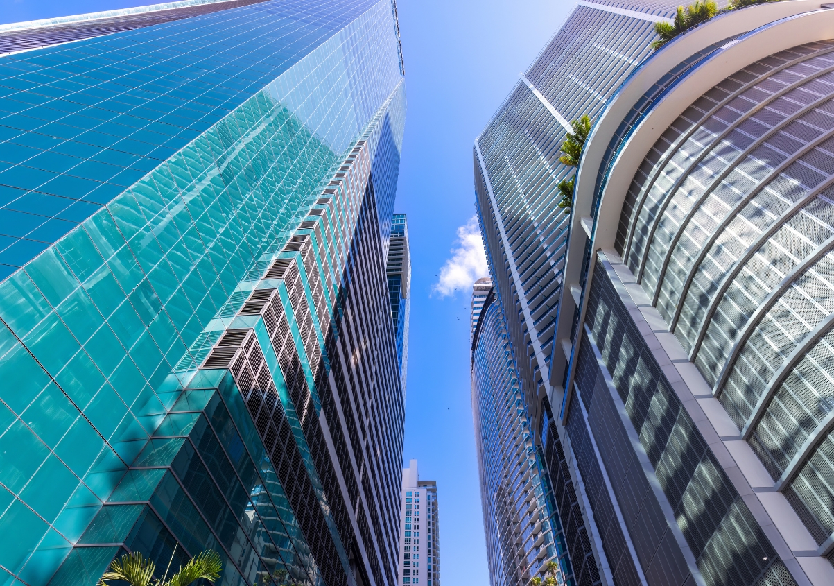 Tall modern skyscrapers with glass facades rise toward a clear blue sky, reflecting sunlight. The buildings have sleek, angular designs and a few small green plants are visible on a terrace.