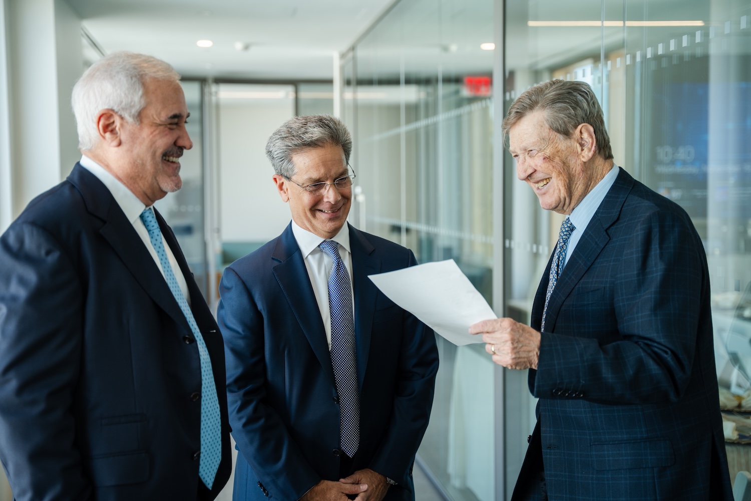 Three older men in business suits stand together in a modern office hallway, smiling and laughing as one of them holds and reads from a sheet of paper. Glass walls and office space are visible in the background.