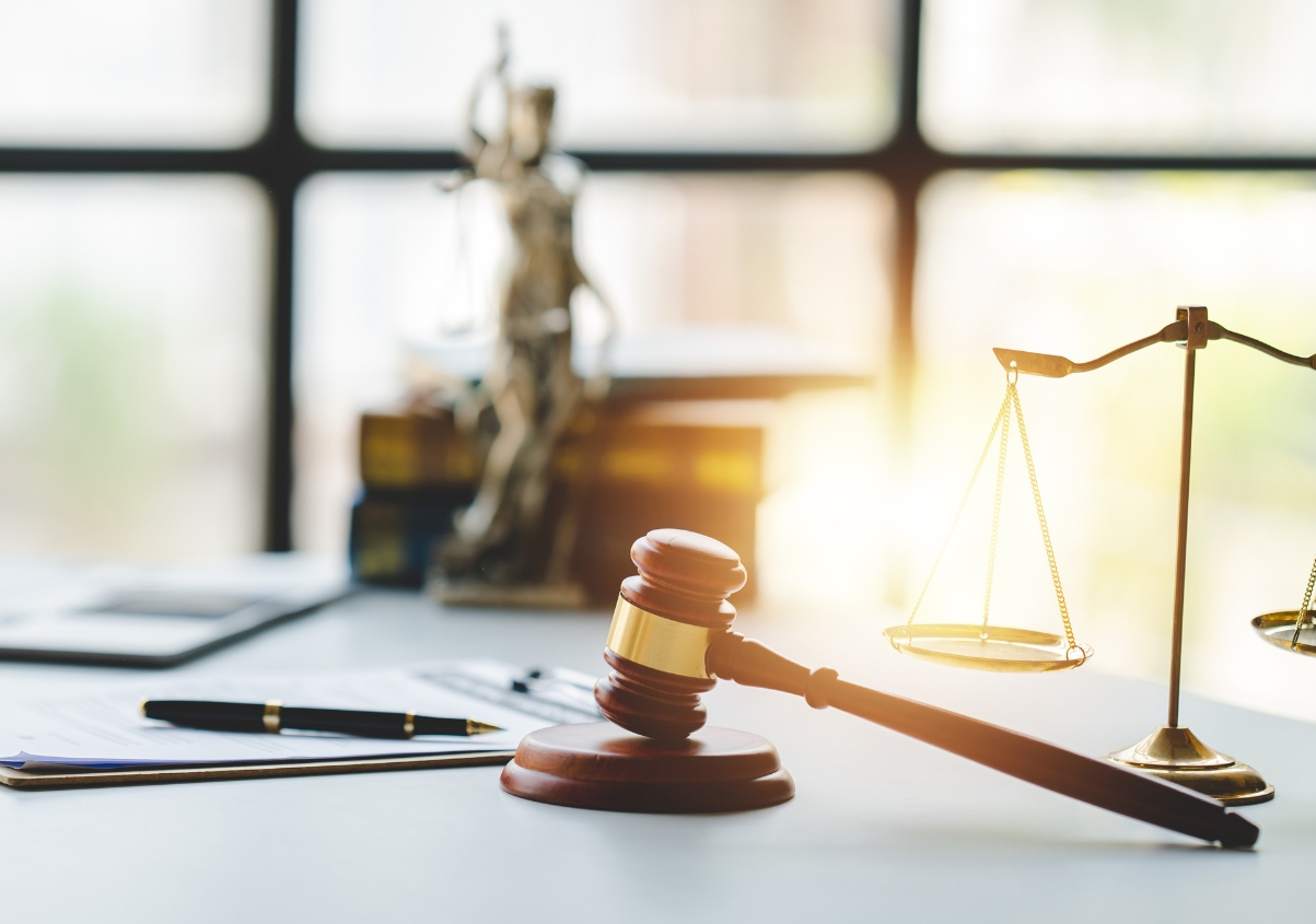 A judge’s gavel, scales of justice, and a pen rest on a desk in a sunlit office, with blurred law books and a Lady Justice statue in the background.