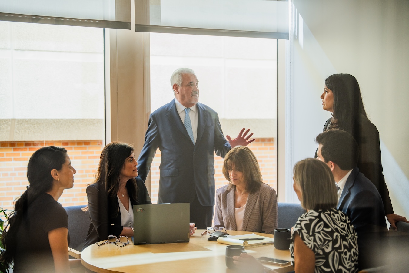 A group of seven business professionals, six seated and one standing, have a discussion around a round conference table with laptops and notebooks in a bright office setting.