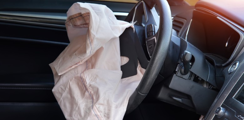 A deployed airbag hangs from the steering wheel inside a car, indicating it has recently been activated, with the interior dashboard and drivers seat partially visible.