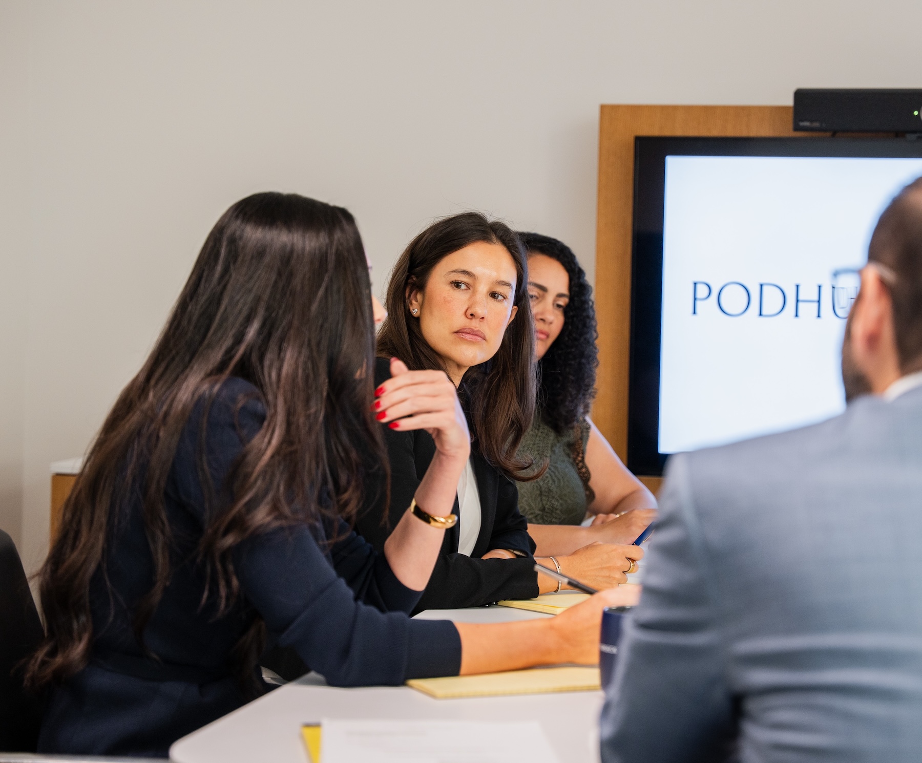 Four people sit at a conference table having a discussion; one woman in the center looks attentively at a colleague. A screen in the background displays the word PODH.