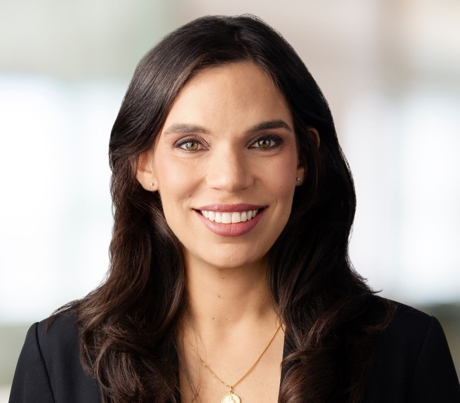 A woman with long dark hair, wearing a black blazer and a gold necklace, smiles at the camera against a blurred light background.