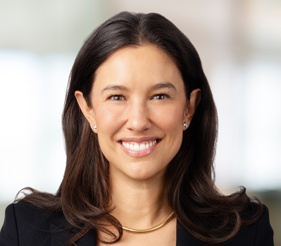 A woman with long brown hair, wearing a black blazer, gold necklace, and stud earrings, smiles at the camera against a softly blurred background.