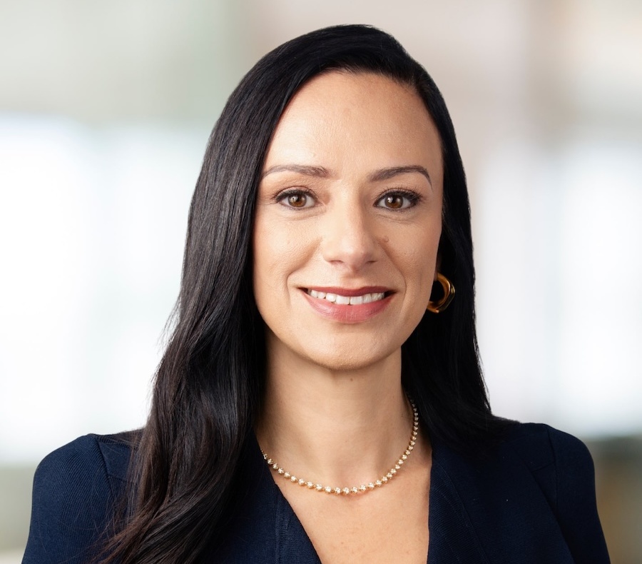 A woman with long dark hair, wearing a navy blue blazer, gold hoop earrings, and a pearl necklace, smiling in front of a blurred light background.