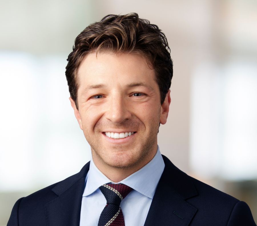 A man with short, wavy brown hair wearing a navy suit, light blue shirt, and patterned tie, smiling at the camera against a blurred, light background.