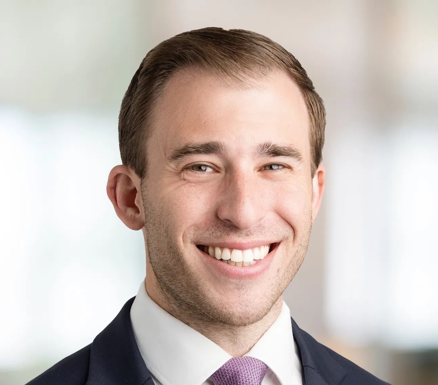 A young man in a navy suit, white shirt, and lavender tie smiles at the camera. He has short brown hair and stands in front of a softly blurred indoor background.